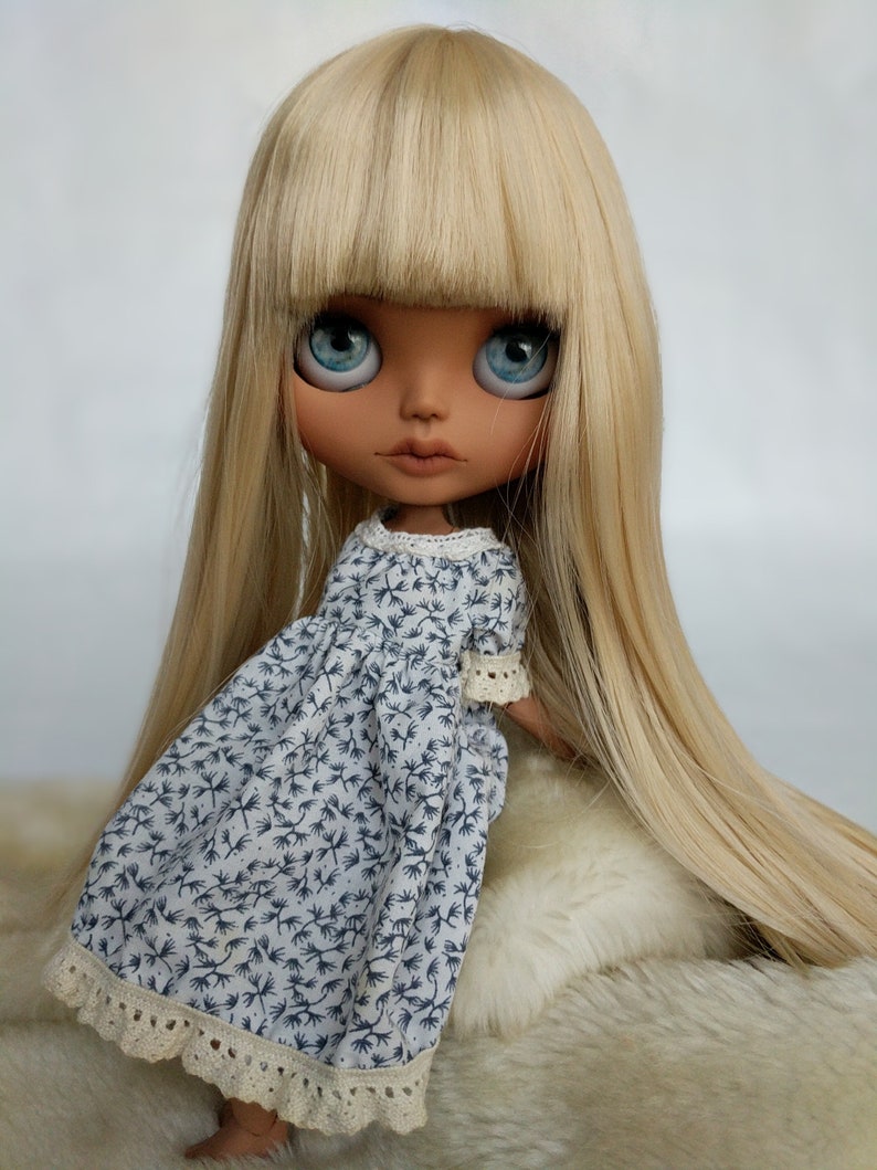 Puede incluir: Una mu&ntilde;eca con el pelo largo y liso rubio y ojos azules que lleva un vestido floral azul y blanco con un borde de encaje.