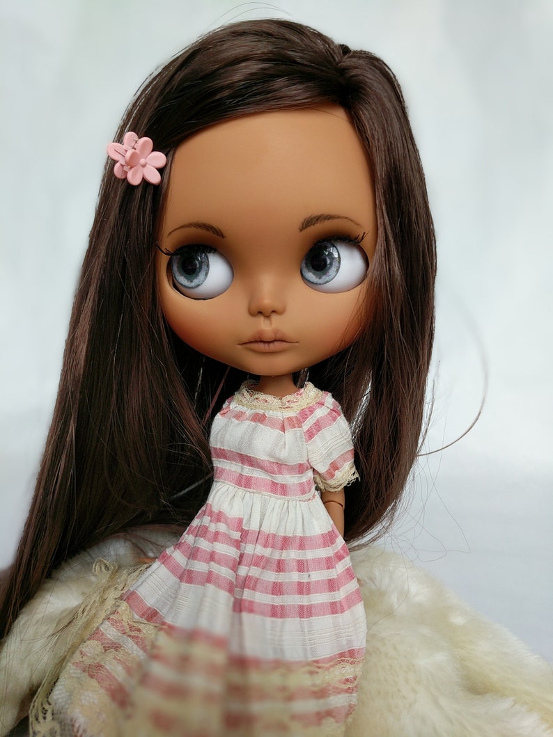 Puede incluir: Una mu&ntilde;eca de piel morena con el pelo largo y casta&ntilde;o, que lleva un vestido a rayas rosa y blanco con ribete de encaje. La mu&ntilde;eca tiene grandes ojos azules y una flor rosa en el pelo.