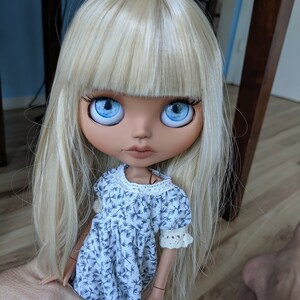 Puede incluir: Una mu&ntilde;eca con el pelo largo y rubio, ojos azules y un vestido blanco con estampado floral.