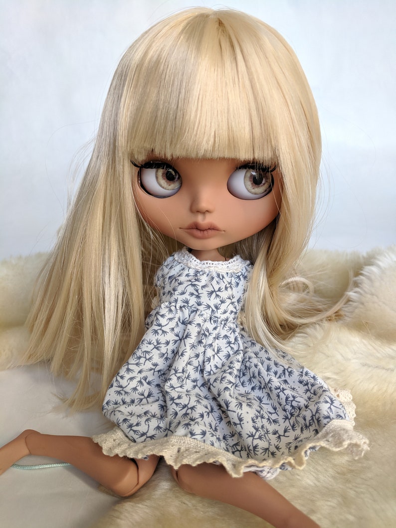 Puede incluir: Una mu&ntilde;eca con el pelo largo y rubio y flequillo que lleva un vestido floral azul y blanco con ribete de encaje.