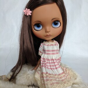 Puede incluir: Una mu&ntilde;eca de piel morena con cabello largo y casta&ntilde;o, ojos azules y una pinza para el pelo con forma de flor rosa. Lleva un vestido a rayas rosa y blanco con ribete de encaje.