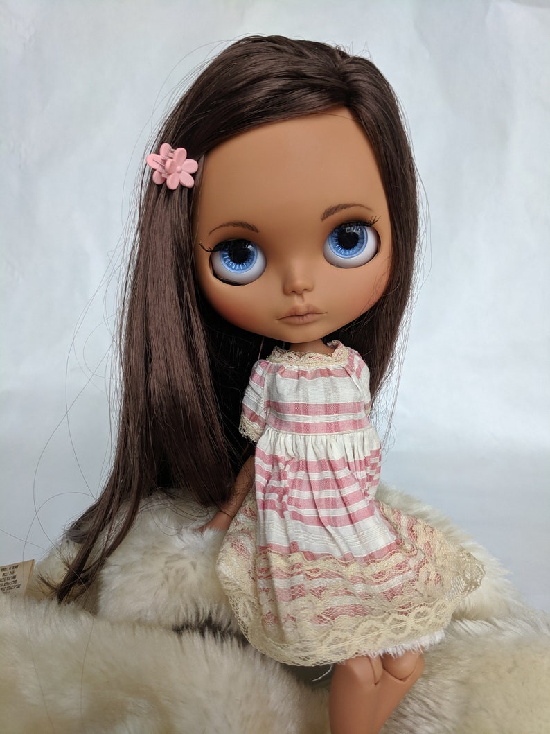 Puede incluir: Una mu&ntilde;eca de piel morena con cabello largo y casta&ntilde;o, ojos azules y una pinza para el pelo con forma de flor rosa. Lleva un vestido a rayas rosa y blanco con ribete de encaje.