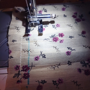 Puede incluir: Primer plano de una aguja de m&aacute;quina de coser y un pie que cose un trozo de tela con un patr&oacute;n floral. La tela es de color beige claro con peque&ntilde;as flores rosas y tallos gris oscuro.