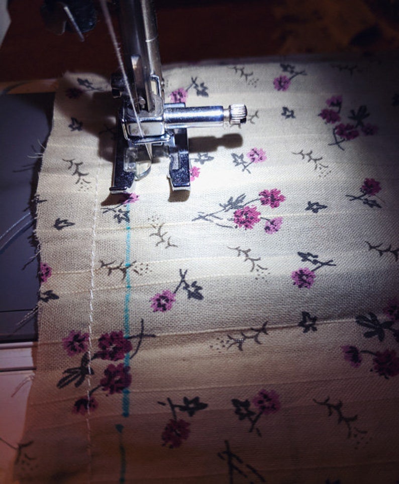 Puede incluir: Primer plano de una aguja de m&aacute;quina de coser y un pie que cose un trozo de tela con un patr&oacute;n floral. La tela es de color beige claro con peque&ntilde;as flores rosas y tallos gris oscuro.
