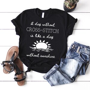May include: A black t-shirt with a white graphic that says "A day without cross-stitch is like a day without sunshine". The graphic also includes a stylised sun and waves.