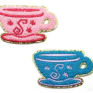 May include: Two embroidered iron-on patches of teacups. One is pink with a gold outline and the other is blue with a gold outline. Both have a white 'S' and white stars.
