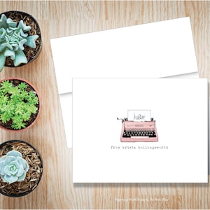 May include: A white greeting card with a pink typewriter illustration and the word "hello". The card is accompanied by a white envelope and three potted succulents. The text "from krista hollingsworth" is below the typewriter.