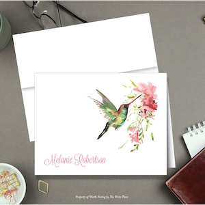 May include: A white card with a watercolor painting of a hummingbird with green and brown feathers, drinking nectar from pink flowers. The card has the name "Melanie Robertson" printed in pink cursive.