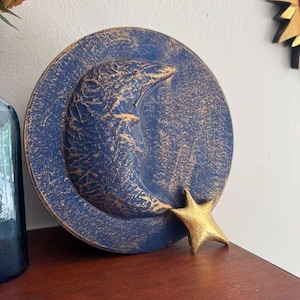 May include: A circular wall plaque featuring a raised crescent moon with a face, finished in a deep blue with distressed gold highlights. A separate, solid gold-colored star is attached to the lower right edge of the circular blue moon base.
