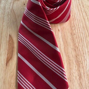 May include: A rolled red tie with white stripes. The tie is made of a smooth fabric and is displayed on a wooden surface. The stripes are evenly spaced and run the length of the tie, creating a classic pattern.