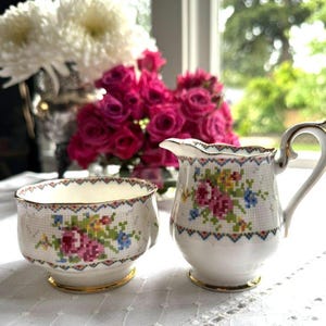 May include: A white porcelain creamer and sugar bowl set featuring a delicate cross-stitch style floral pattern in shades of pink, blue, yellow, and green. Both pieces have a scalloped rim with a geometric border and gold-toned trim.