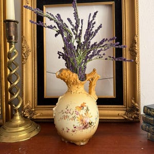 May include: A cream-colored ceramic pitcher with a rounded body and an ornate handle, featuring a painted floral design of yellow and pink flowers. The pitcher is filled with a bouquet of purple lavender-like sprigs, showcasing a textured rim.