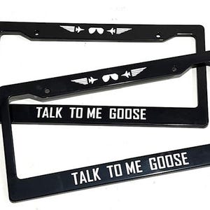 May include: Black license plate frames with the text "TALK TO ME GOOSE" in white. The top of the frames feature a white graphic of aviator sunglasses and wings. The frames are rectangular and have a glossy finish.
