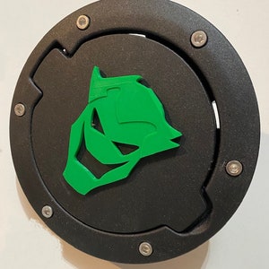 May include: A black round gas cap cover with a green stylized animal head design. The cover has six silver screws around the edge.