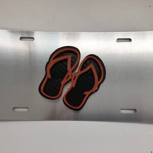 Könnte beinhalten: Ein silbernes Nummernschild aus Metall mit zwei schwarzen Flip-Flops mit orangefarbenen Umrissen.