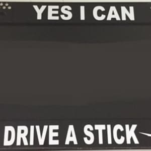 May include: Black license plate frame with rhinestones around the border. The frame has white text that reads "YES I CAN DRIVE A STICK" with a silhouette of a witch flying on a broomstick.