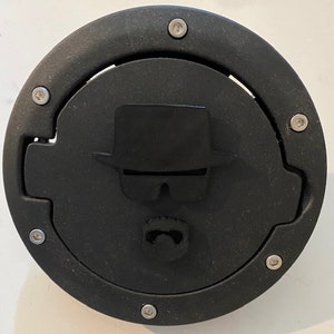 Può includere: Tappi di benzina neri con la sagoma di un uomo con cappello e occhiali da sole. L'uomo ha baffi e barba.