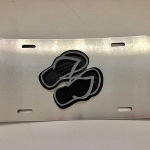 Könnte beinhalten: Ein silbernes Nummernschild aus Metall mit einer schwarzen Silhouette von zwei Flip-Flops.