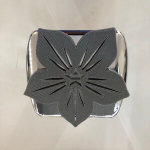Può includere: Un porta fazzoletti di colore grigio, stampato in 3D, a forma di fiore con un ritaglio a forma di stella al centro. Il supporto è montato su una base argentata.