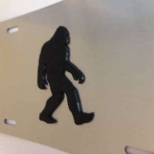 May include: A black silhouette of Bigfoot walking on a silver metal license plate.