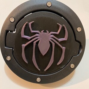 Può includere: Un tappo serbatoio rotondo nero con un designo a forma di ragno viola. Il tappo serbatoio ha un coperchio incernierato ed è fissato con viti.