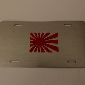 May include: A silver metal license plate with a red rising sun design.