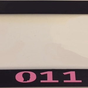 May include: Black license plate frame with a pink silhouette of a person and the number 011.
