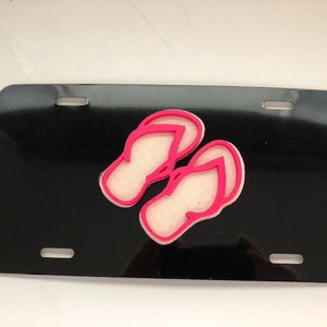 Könnte beinhalten: Schwarzes Nummernschild mit einem Paar rosa und weißen Flip-Flops.
