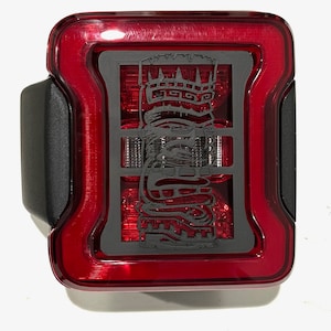 Puede incluir: Luz trasera roja con un diseño tribal negro superpuesto. La luz trasera es rectangular con esquinas redondeadas y tiene una carcasa de plástico negro.