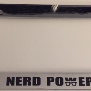 May include: A shiny chrome license plate frame with the words "NERD POWER" in black block letters. The word "POWER" includes a graphic of eyeglasses. The frame has a rectangular shape.