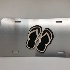 Könnte beinhalten: Ein silbernes Nummernschild aus Metall mit einem schwarzen und weißen Ausschnitt von zwei Flip-Flops.