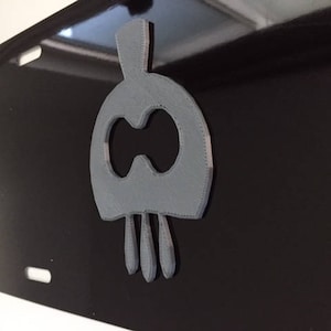 Puede incluir: Un diseño de calavera gris impreso en 3D con un agujero en el centro y tres puntas en la parte inferior. La calavera está unida a una superficie negra.