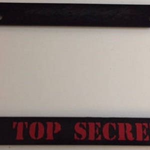 Könnte beinhalten: Schwarzer Kennzeichenrahmen mit dem roten Schriftzug "TOP SECRET". Der Rahmen hat eine rechteckige Form mit abgerundeten Ecken und Befestigungslöchern oben.