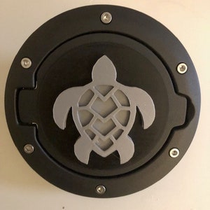 May include: A black, circular object with a silver turtle design in the center. The turtle is a stylized silhouette. The object is secured with six silver screws around the perimeter. The background is a light beige color.