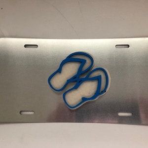 Könnte beinhalten: Ein silbernes Nummernschild aus Metall mit zwei blauen Flip-Flops in der Mitte.