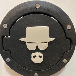 Può includere: Un tappo di benzina rotondo nero con una silhouette bianca di una persona che indossa un cappello, occhiali da sole e baffi.