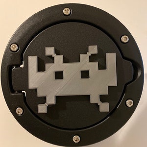 Può includere: Un tappo del serbatoio nero con una grafica aliena a 8 bit argentata. Il tappo del serbatoio ha un design circolare con un bordo rialzato e quattro viti argentate attorno al bordo.