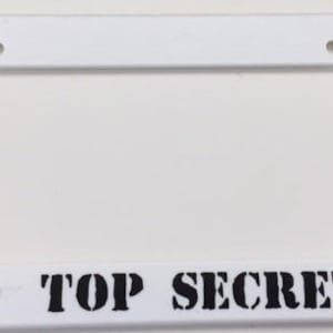 Può includere: Telaio per targa in plastica bianca con testo nero che recita "TOP SECRET".