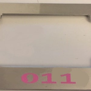 May include: Chrome license plate frame with a pink "011" printed on the bottom.