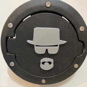 Può includere: Un tappo serbatoio rotondo nero con un design 3D argentato di una persona con cappello, occhiali da sole e barba.