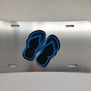 Könnte beinhalten: Ein silbernes Nummernschild aus Metall mit zwei schwarzen Flip-Flops mit blauen Umrissen.