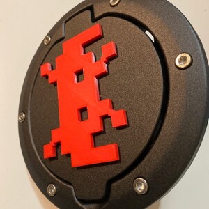 Può includere: Un tappo serbatoio rotondo nero con un design alieno pixelato rosso. Il tappo serbatoio ha una superficie testurizzata e un anello argentato attorno al bordo.