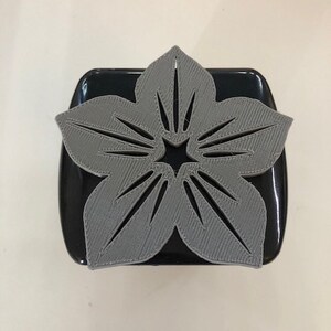 Può includere: Un fiore grigio stampato in 3D con un centro a forma di stella. Il fiore è su una superficie nera.