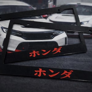 Puede incluir: Marcos negros para matrículas con caracteres japoneses rojos, apilados frente a un coche blanco con detalles rojos. Los marcos tienen forma rectangular con orificios de montaje.