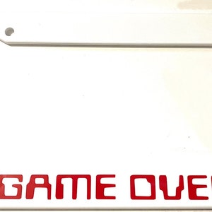 May include: White plastic license plate frame with red text that reads "GAME OVER".