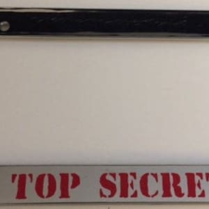 Peut inclure: Un cadre de plaque d'immatriculation chromé avec les mots "TOP SECRET" en lettres rouges. Le cadre a une surface brillante et réfléchissante et une barre supérieure noire avec deux petites vis.