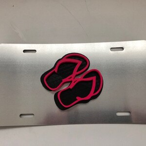 Könnte beinhalten: Ein silbernes Nummernschild aus Metall mit einem schwarzen und pinkfarbenen Flip-Flop-Design.