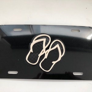 Könnte beinhalten: Schwarzes Nummernschild mit einem weißen Umriss von zwei Flip-Flops.