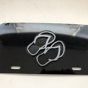 Könnte beinhalten: Schwarzes Nummernschild mit einem silbernen 3D-Design von zwei Flip-Flops.