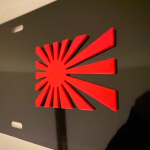 May include: Black license plate featuring a raised red sunburst design. The sunburst has a central red circle with radiating red lines. The plate has four mounting holes.